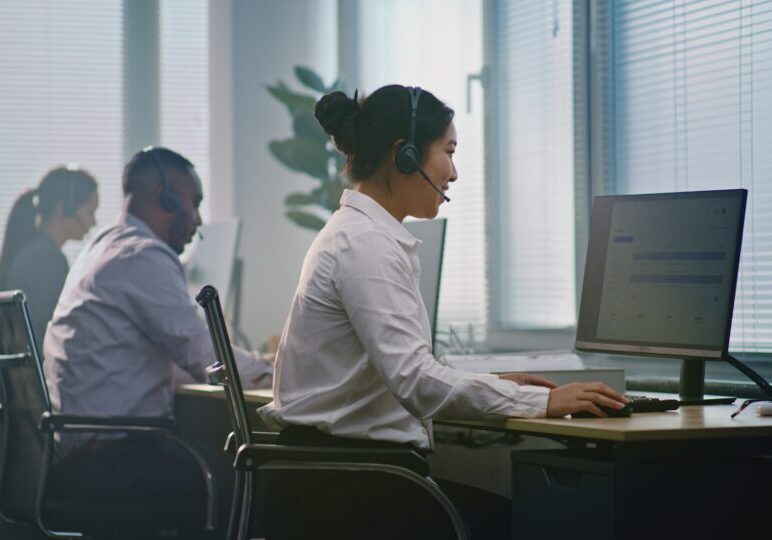 Asian female helpdesk specialist talks on headset, uses computer, looks at camera