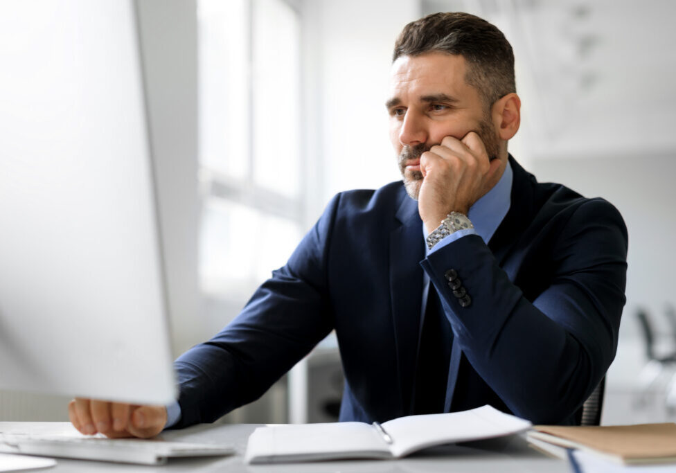 Bored middle aged businessman sitting in front of computer at workplace in office, leaning head on hand and looking at screen. Upset man suffering job problems, reading bad negative news